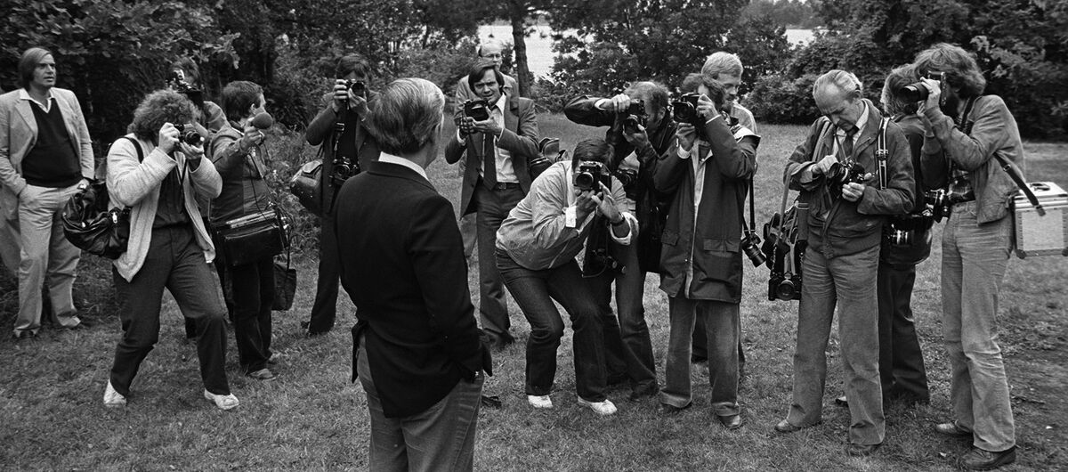 Schwarz-weiß Foto aus den 70er Jahren, auf dem Helmut Schmidt auf einer Wiese mit Blick auf einen See steht. Er ist von rund 12 Fotografen umringt. Das Foto ist hinter ihm aufgenommen worden.