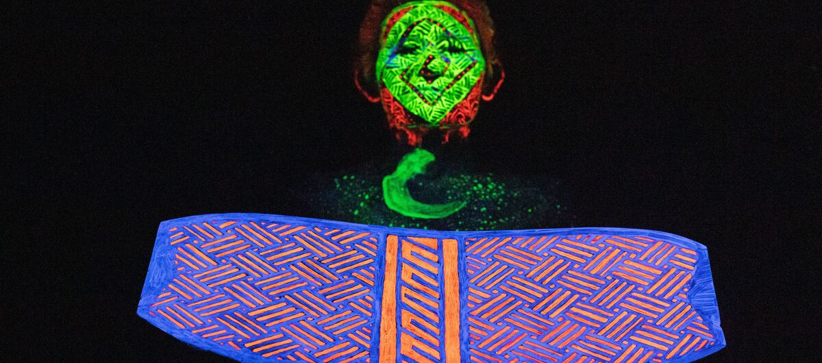 Das Bild zeigt im Vordergrund eine Art Schild, in das kunstvoll ein systematisches Muster aus Strichen geschnitzt ist - mit blauer und orangener Farbe bemalt. Dahinter ein Mensch, dessen Gesicht und Brust mit fluoreszierender Farbe bemalt wurde.
