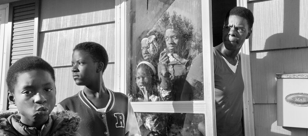 Schwarz-weiß Foto in Nahaufnahme vor einem Haus. Zwei Jugendliche stehen vor der Haustür. Eine Frau steht zwischen Haustür und Fliegengitter und schaut heraus. Drei Jugendliche stehen hinter dem Fliegengitter. Alle Menschen haben eine dunkele Haut.