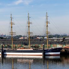 Der Frachtsegler Peking liegt im Hafen vor Anker. Es ist ein vier Masten tragendes Schiff mit insgesamt 32 Segeln, die eingeholt sind. Rechts und links des Schiffes steht je ein Hafenkran. Es ist ein freundlicher Tag.