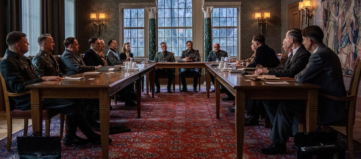 Filmausschnitt aus dem Film "Die Wannsee-Konferenz". An u-förmig gestellten Holztischen in einem Raum mit großen Fenstern und verzierten Wänden sitzen 13 Männer und zwei Frauen. Alle tragen Anzug. Es liegen Papiere und Mappen auf den Tischen.