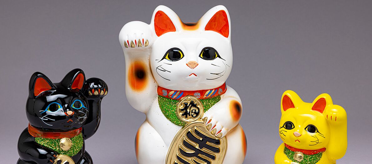 Auf dem Bild sind drei Maneki-Neko, die japanischen Glückskatzen, zu sehen. Sie unterscheiden sich in Größe und Farbe. Jede Katze hat ein freundliches Gesicht und hält eine Pfote erhoben, wie es typisch für Maneki-Neko ist.