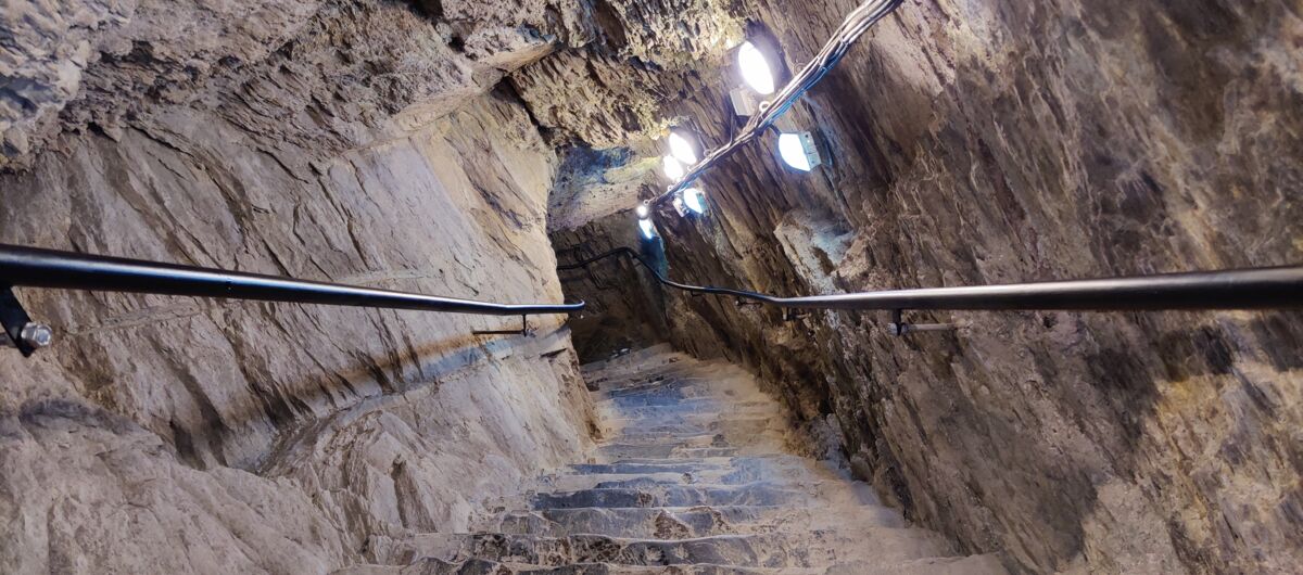 Foto von einer Steintreppe, die nach unten in einen Fels führt. Rechts und link der Treppe sind schwarze Handläufe an die Felsfände angebracht. Auf der rechten Seite sind leuchtende Lampen an die Wand angebracht.