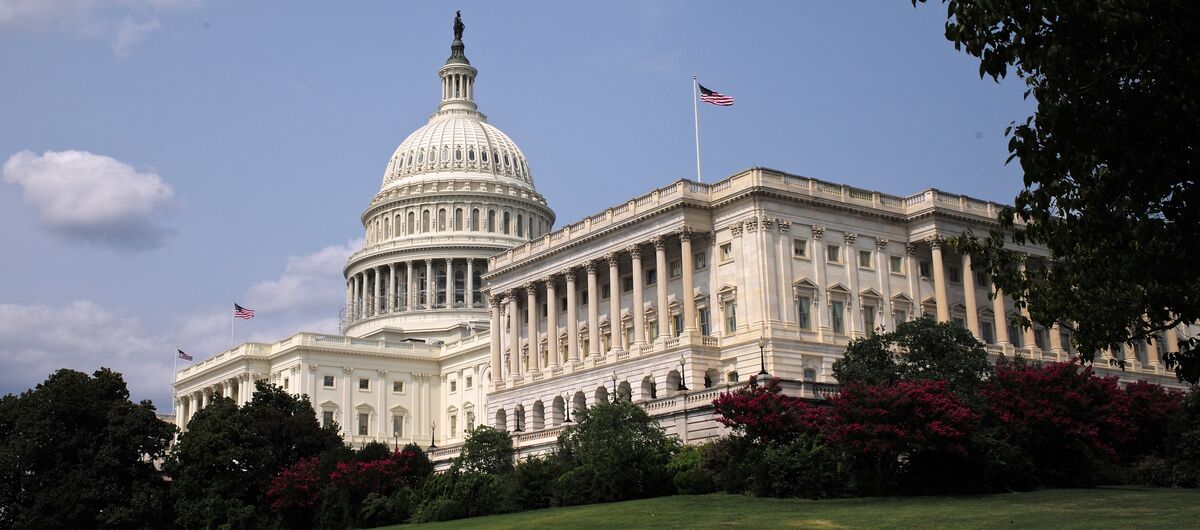 Das Kapitol in Washington D. C. mit seiner großen weißen Kuppel und der amerikanischen Flagge auf dem Dach. Im Vordergrund liegt eine grüne Rasenfläche mit Bäumen und Sträuchern, die das Regierungsgebäude einrahmen.