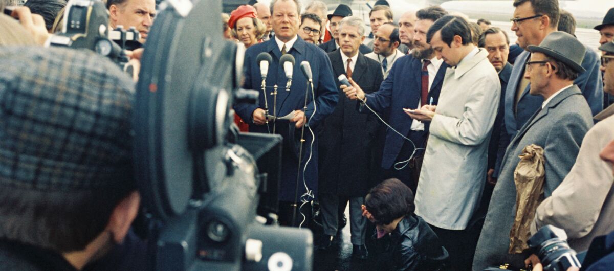 Willy Brandt steht draußen vor Mikrofonen. In der Hand hält er einen Zettel. Er spricht. Um ihn scharren sich Menschen, die teils Handmikrofone zu ihm und Videokameras auf ihn richten.