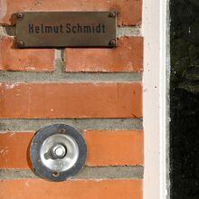 Foto eines kleinen Ausschnittes einer Hauswand aus Backstein, in die eine runde Klingel mit Klingeknopf eingelassen sowie ein Metalschild befestigt ist. Auf dem Schild steht "Helmut Schmidt".