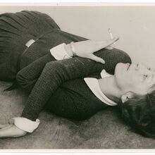 Schwarz-weiß Foto einer liegenden Frau, die von Kopf bis Oberschenkel zu sehen ist. Ihr rechter Arm liegt quer über der Brust, die Hand berührt den Boden. Der linke Arm ist angewinkelt. Die Hand hält eine Zigarette und liegt auf dem rechten Oberarm.