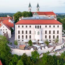 Luftfoto des Diözesanmuseum Freising. Auf einem Berg steht das viergeschossige Haus. Davor ist ein Platz, auf dem Sonnenschirme stehen und der halbrund eingemauert ist. grüne Bäume schmiegen sich dicht hangabwärts an diesen Komplex.