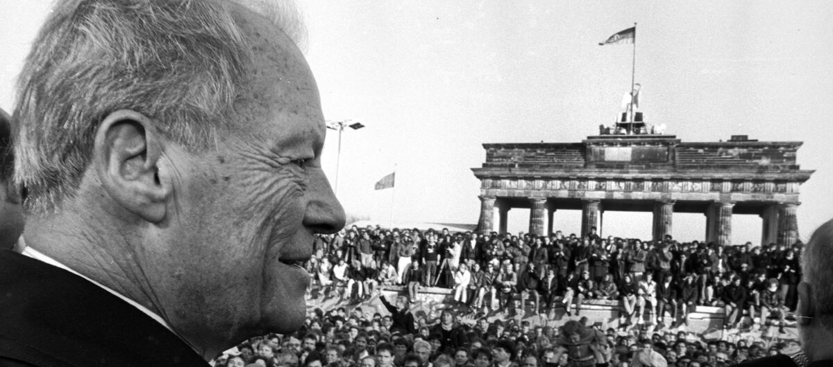 Schwarz-weiß Fotografie von Willy Brandt, der links im Bildrand steht. Er blickt auf eine Masse an Menschen, die sich vor dem Brandenburger Tor, welches im Hintergrund zu sehen ist, versammelt haben.