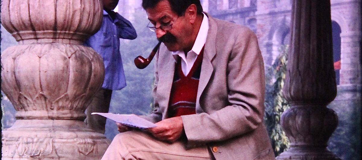 Foto von Günter Grass in Delhi. Er sitzt auf einer Treppe. Seine Beine hat er überschlagen. Er hat eine Pfeife im Mund. In den Händen hält er ein Papier, auf das er blickt. Neben ihm steht eine schwarze Tasche. Im Hintergrund steht ein indischer Junge.