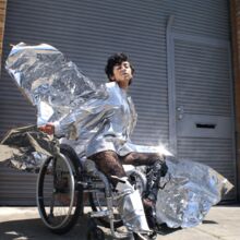 Eine Frau posiert im Rollstuhl. Sie trägt ein silber-glänzendes kurzes Kleid mit langen Ärmeln. Ein Bein steckt in einem silbernen Stiefel, das andere umfasst eine Art Prothese. Hinter ihr weht ein silberner Umhang, der im Licht glitzert.