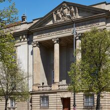 Foto der Außenansicht der Sammlung Schack der Bayerischen Staatsgemäldesammlungen. Der historische Galeriebau wird leicht durch Bäume verdeckt. Die Sonne scheint.