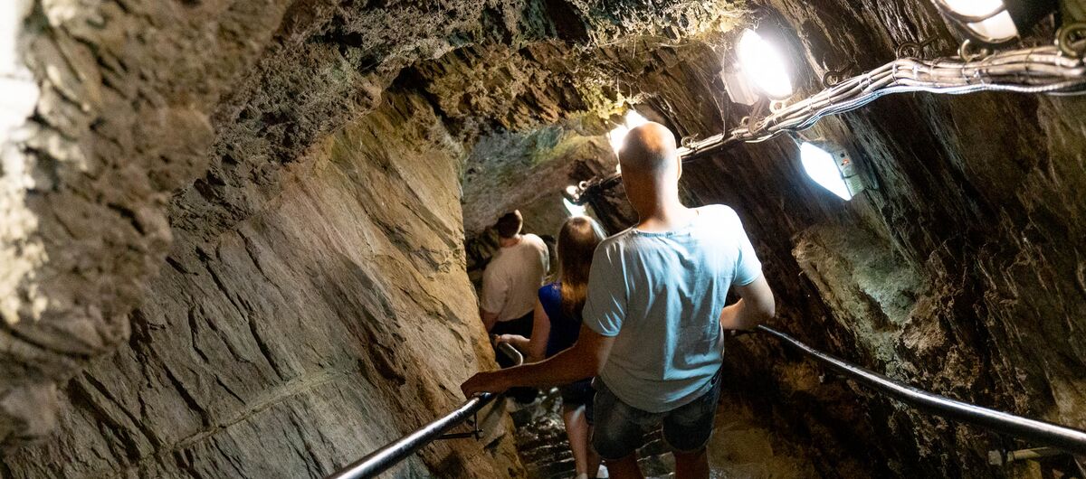 Foto von einer Steintreppe, die nach unten in einen Fels führt. Rechts und link der Treppe sind schwarze Handläufe an die Felsfände angebracht. Auf der rechten Seite sind leuchtende Lampen an die Wand angebracht. Drei Personen gehen die Treppe hinab.