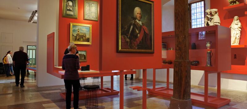 Blick in Ausstellungsraum mit Boden aus Steinplatten in verschiedenen Farben und Größen. An roten mobilen Wänden und in Vitrinen werden Gemälde, Skulpturen und andere Dinge ausgestellt. Fünf Personen schauen sich die Ausstellung an.