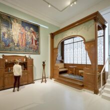 Raum im Museum, in dem eine Frau vor einem Holzvertiko steht, welches kunstvoll geschnitzte Türen aufweist. Darüber hängt ein übergroßes Bild mit biblischen Inhalten. Daneben befindet sich ein Nachbau mit Sitzmöbeln, Fenstern und hölzernem Rundbogen.