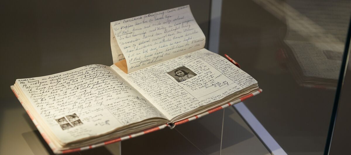 Foto einer originalgetreuen Kopie des Tagebuchs von Anne Frank. Das Buch liegt aufgeschlagen in einer Vitrine. Auf der linken Buchseite sind vier kleine Portraitfotos in den Text gelebt. Auf der rechten Seite ein größeres.