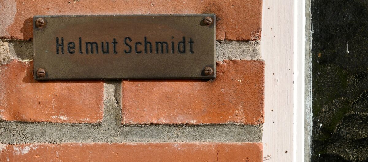 Foto eines kleinen Ausschnittes einer Hauswand aus Backstein, in die eine runde Klingel mit Klingeknopf eingelassen sowie ein Metalschild befestigt ist. Auf dem Schild steht "Helmut Schmidt".