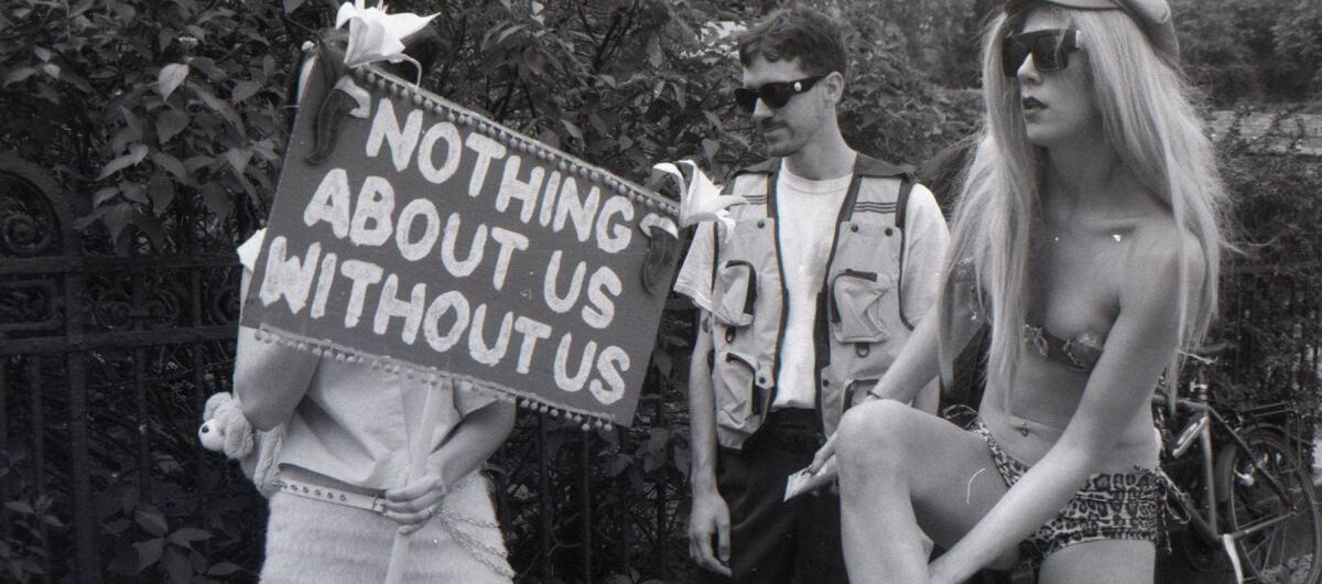 Schwarz-weiß Foto mit drei Personen im Freien. Die linke Person scheint eine Frau zu sein. Sie hält ein Schild mit der Aufschrift "Nothig about us without us" vor ihr Gesicht. Mittig steht ein Mann. Rechts eine Frau im Bikini, die sich ein Bein eincremt.