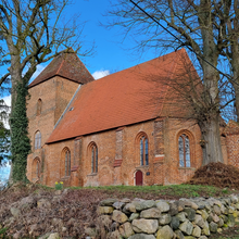 Foto der Außenansicht der Dorfkirche Friedrichshagen, die leicht erhöht steht und von einer Natursteinmauer umgebene ist. Die Kirche besteht aus roten Ziegeln und hat ein rotes Dach. Drumherum stehen hohe unbelaubte Bäume.