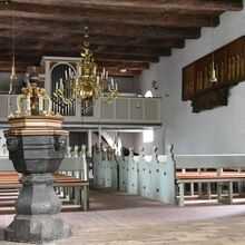 Foto des Kirchenraums. Im Vordergrund ist das Taufbecken zu sehen. Dahinter sind die Kirchbänke in zwei Reihen nebeneinander aufgestellt - oberhalb dazwischen ein Kronleuchter. Im Hintergrund ist die Orgel zu sehen. Diese Decke besteht aus Holzbalken.