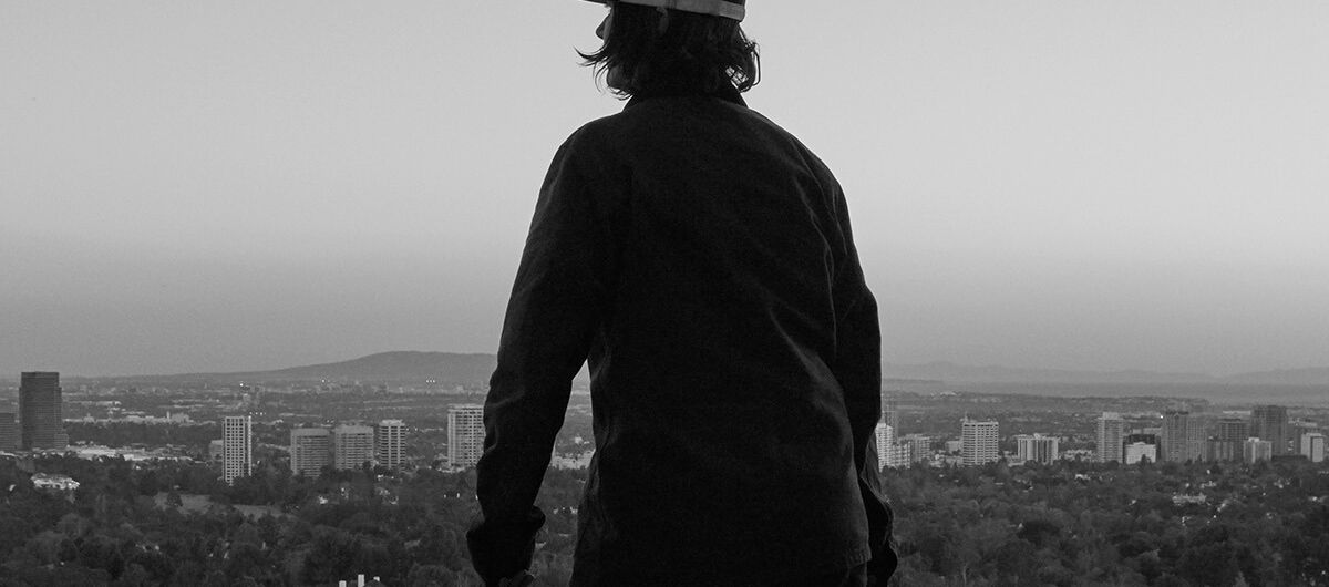 Schwarz-weiß Foto. Ein Mensch, der von hinten zu sehen ist, steht am Rande einer Plattform und hat in der rechten Hand einen kleinen Koffer. Hinter dieser Szene liegen tief unten Wald und Stadt mit Hochhäusern. Der Ausblick ist weit.