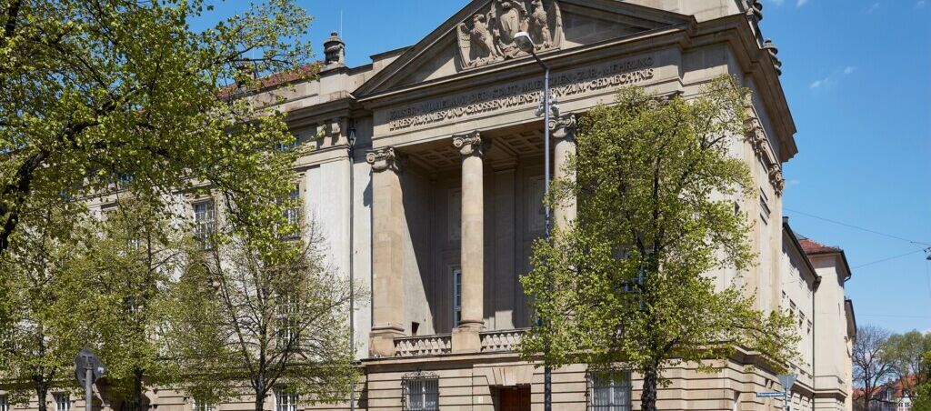 Foto der Außenansicht der Sammlung Schack der Bayerischen Staatsgemäldesammlungen. Der historische Galeriebau wird leicht durch Bäume verdeckt. Die Sonne scheint.