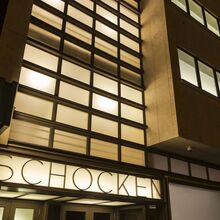 Foto einer mehrstöckigen Gebäudefront bei Nacht. Viele moderne Fenster zieren die Front. Im Inneren des Gebäudes brennt Licht. Über dem Eingang steht der Schriftzug "Schocken".