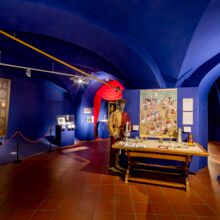 Blick in Ausstellungsraum mit royalblauen Wänden und Decken sowie braun gefliestem Boden. Die Decken sind bogenartig geschwungen. Ausgestellt werden an lebensgroßen Puppen Kleidungsstücke sowie Karten und Alltagsgegenstände.