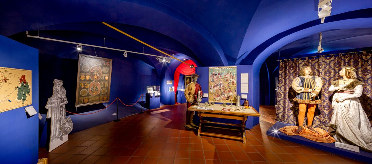 Blick in Ausstellungsraum mit royalblauen Wänden und Decken sowie braun gefliestem Boden. Die Decken sind bogenartig geschwungen. Ausgestellt werden an lebensgroßen Puppen Kleidungsstücke sowie Karten und Alltagsgegenstände.