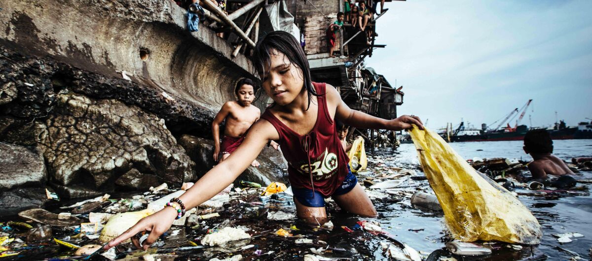 Das Foto zeigt im Vordergrund ein Kind mit leicht dunkler Haut, das kniehoch im vermüllten Wasser steht. Es streckt den Arm aus und sammelt Müll in einer Plastiktüte. Im Hintergrund sind weitere Kinder im und am Wasser zu sehen.