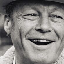Schwarzweiß Fotoportrait von Willy Brandt, lachend, seine Haare unter einem Hut versteckt, dessen Krempe man sieht. Das Lachen legt die obere Zahnreihe frei, in der ein Zahn glänzend hervorscheint. Die Augen sind von Lachfalten gezeichnet.