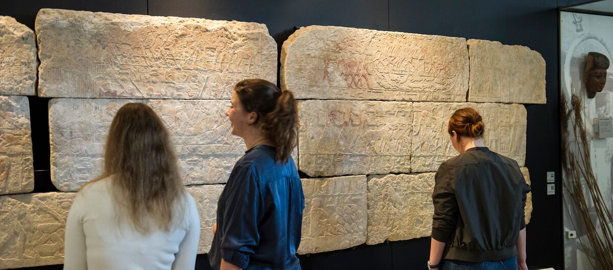 Foto von drei Menschen, die vor einer schwarzen Wand mit hellen großen Steinplatten stehen. In diesen sind verschiedene Szenen großflächig hineingemeißelt. Die Steinplatten haben ein Querformat und sind in drei Reihen übereinander angeordnet.