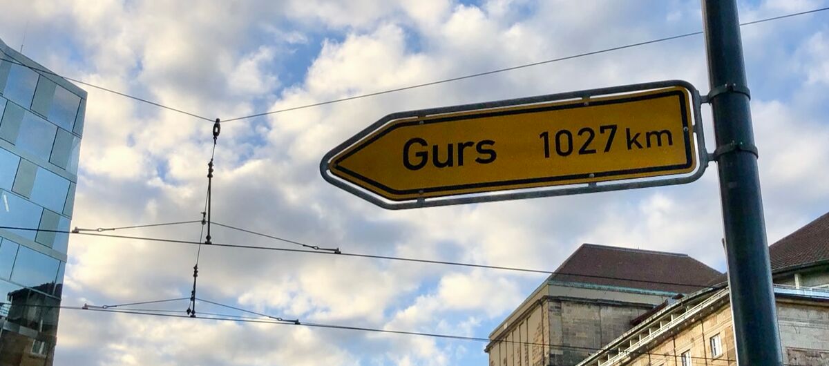 Foto eines gelben Straßenschildes, auf dem Gurs 1027 km steht. Es zeigt nach links. Im Hintergrund sind Teile von älteren und neueren Gebäuden sowie Oberleitungen von Straßenbahnen zu sehen.