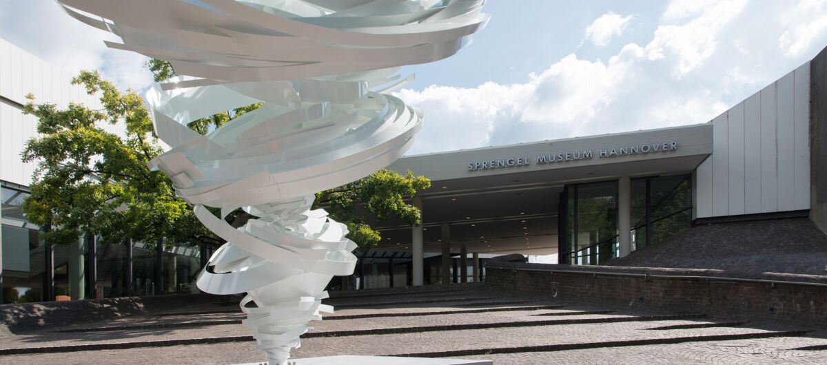 Außenansicht des Sprengel Museums. Vor dem Eingang steht eine Skulptur, die einen Wirbelsturm, eine Tornado-Plastik zeigt.