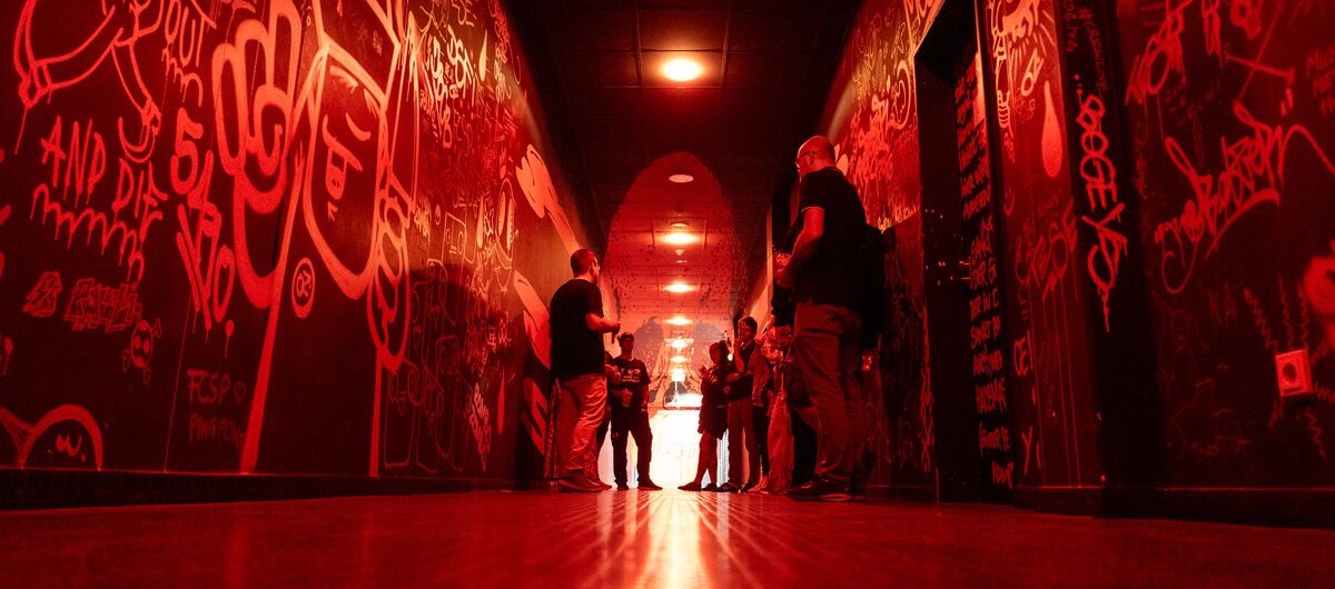 Foto eines langen Ganges, dessen Wände mit Graffiti besprüht sind. Das Foto ist vom Fußboden aus aufgenommen. Der Flur ist in rotes Licht getaucht. In der Mitte des Flures stehen Menschen.