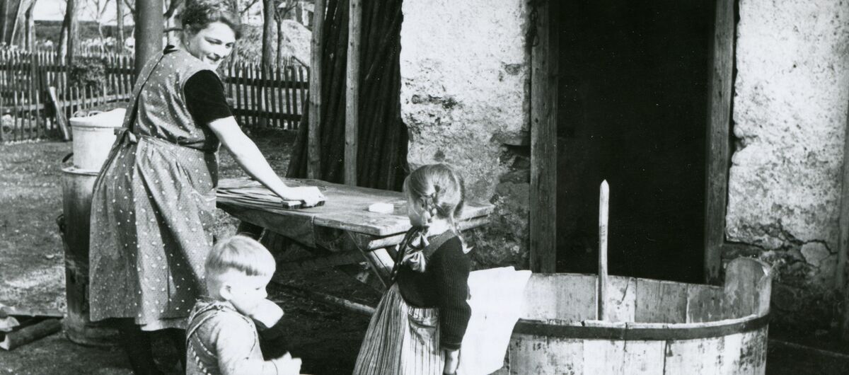 Schwarz-weiß Foto einer Alltagsszene einer Familie. Eine Frau arbeitet mit einem Mädchen und einem jüngeren Jungen vor einem Haus. Die Frau schaut freundlich über die Schulter zu den Kindern.