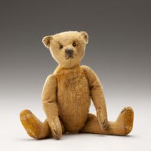 Ein hellbrauner Teddybär sitzt auf einem weißen Untergrund. Die Unterseiten der Hintertatzen sind mit weißen Punkten und Strichen gemustert. Leichte Falten und Abnutzungsspuren sind am Teddy zu erkennen.