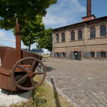 Foto der Metallgießerei des Stadtmuseums Kiel. Das Gebäude besteht aus Stein und hat sechs große Fenster im Obergeschoss sowie vier kleine Fenster und eine Tür im Erdgeschoss. Der Hof vor dem Haus besteht aus Kopfsteinpflaster.