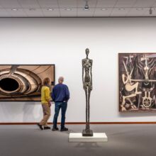 Blick in Ausstellungsraum mit grauem Boden und weißer Wand. Zwei große rechteckige Gemälde hängen an der Wand. Sie sind in Schwarz-, Weiß- und Brauntönen gehalten. Zwischen ihnen steht in den Raum gerückt eine sehr hohe dünne Skulptur.