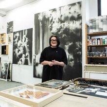 Foto der Künstlerin Lucia Tallová im Atelier. Sie steht hinter einem großen Arbeitstisch und lacht. Rechts hinter ihr ist ein Holzregal mit Kunstmaterialien. Links hinter ihr hängen und stehen schwarz-weiß Bilder in unterschiedlichen Größen.