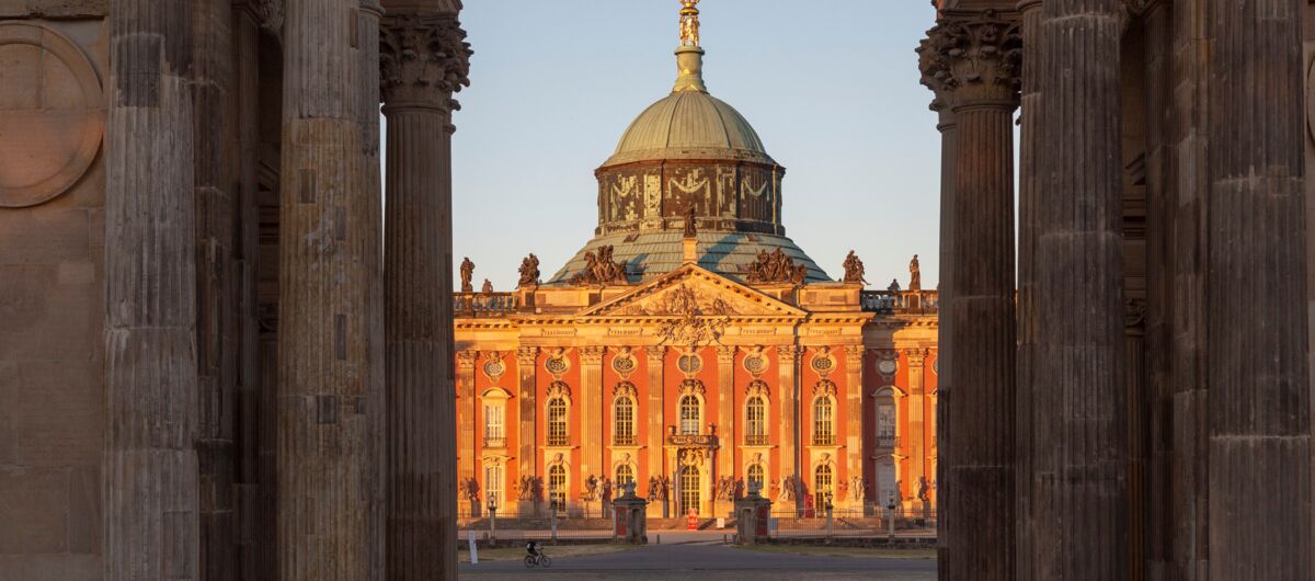 Foto durch einen Durchgang von Steinsäulen auf das Neue Palais, das durch die Abendsonne angeleuchtet wird. Das mehrstöckige verzierte Gebäude hat eine runde Kuppel. Es ist aus Stein. Manche Teile sind mit braun-roter Farbe gestrichen.