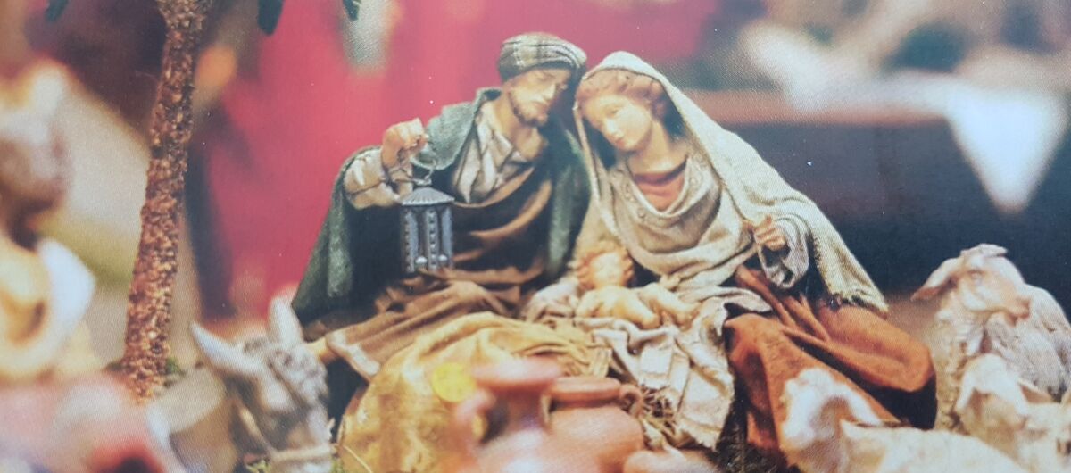 Nahaufnahme von Krippenfiguren. Zusehen sind Maria und Joseph mit dem Jesuskind. Im Vordergrund sind verschwommen drei Schafe auf der linken Seite, in der Mitte vier Tonkrüge und auf der rechten Seite ein Esel zu sehen. Die Figuren sind sehr filigran.