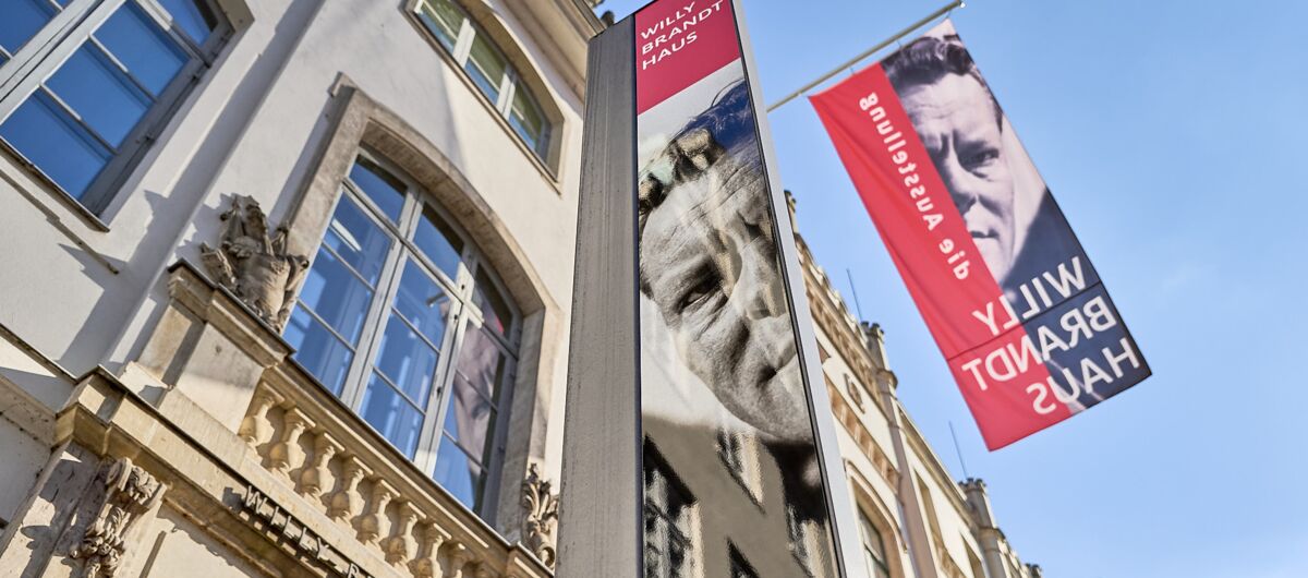 Foto der Außenansicht des Willy-Brandt-Hauses Lübeck. Die Fassade, die mit einer langen und einer kurzen Fahnen des Ausstellungshauses bestückt ist, wurde von unten schräg nach oben fotografiert. Das Haus besteht aus Sandstein.