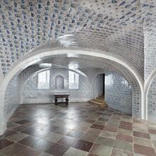 Gewölberaum, der von oben bis unten mit Fliesen ausgekleidet ist. Zwei Fenster sind in Nischen eingelassen. Durch die blau-weißen Fliesen ist der Raum hell, wirkt aber kühl. Ein kleiner dunkler Tisch steht an einer Wand. Stufen führen rechts aus dem Raum.