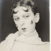 Portraitfoto in schwarz-weiß. Die Person hat kurze Haare, an der Stirn zu zwei kleinen Hörnern geformt. Das Gesicht ist geschminkt, auf den Wangen ist je ein Herz. Tuch und Oberteil sind weiß. Auf dem Oberteil steht: I am in training. Don't kiss me.
