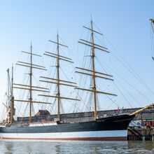 Der Frachtsegler Peking liegt im Hafen vor Anker. Es ist ein vier Masten tragendes Schiff mit insgesamt 32 Segeln, die eingeholt sind. Rechts und links des Schiffes steht je ein Hafenkran. Es ist ein freundlicher Tag.