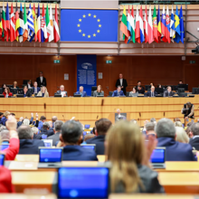 Foto aus dem Plenarsaal des Europäischen Parlaments. Zentral im Hintergrund sind die Vorsitzenden zu sehen. Über ihnen hängt das EU-Logo sowie rechts und links davon die Fahnen der Nationen. Im Vordergrund die Plenumsteilnehmenden von hinten.
