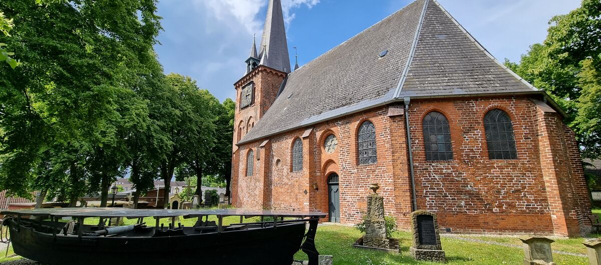 Foto der Fischerkirche St. Andreas in Lübeck. Die Backsteinkirche ist umgeben von Rasenfläche, auf denen historische Grabsteine stehen, und hohen Bäumen. Im Vordergrund der Kirche steht auf einer Fläche mit kleinen Kieselsteinen ein schwarzes Fischerboot.