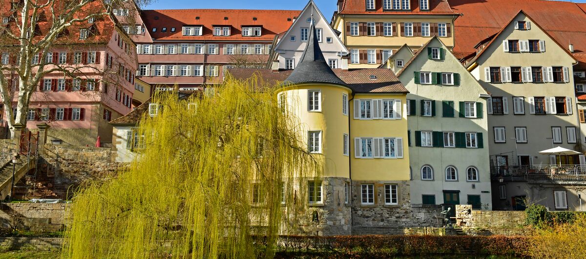 Foto des Hölderlinturms von Seiten des Fluss Neckar. Hinter dem runden Turm mit spitzem Dach schließt sich ein Gebäude an. Die unterste Etage des Gebäudekomplexes ist aus Naturstein, die Fassade der oberen beiden Etagen gelb gestrichen.