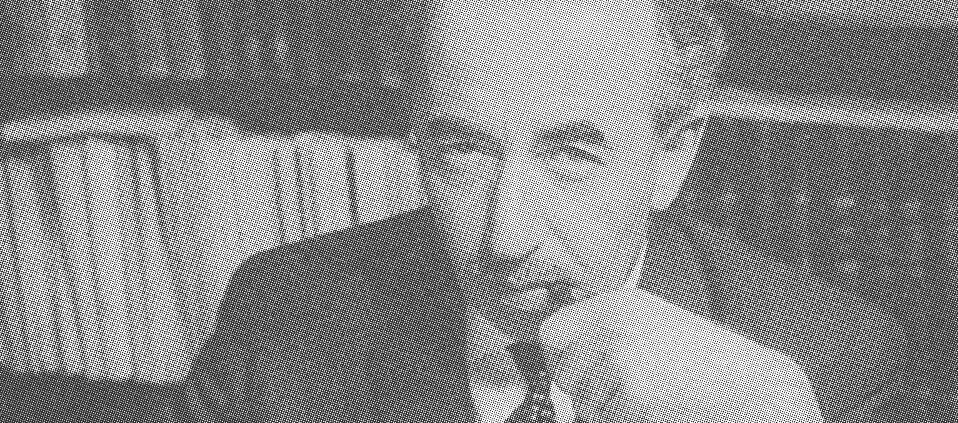 Schwarz-weiß Foto von Joseph Wulf. Er sitzt an einem Tisch, vor ihm Papiere. In der rechten Hand hält er einen Stift. Der linke Arm ist aufgestützt. Die Hand hält eine Pfeife, die zwischen seinen geschlossenen Lippen liegt. Im Hintergrund ein Bücherregal.